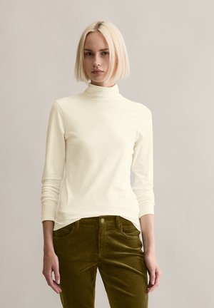 Cremfarbener Rollkragenpullover mit langen Ärmeln aus weichem Stoff, kombiniert mit olivgrünen Cordhosen. Verfügt über ein tailliertes Design und gerippte Textur.