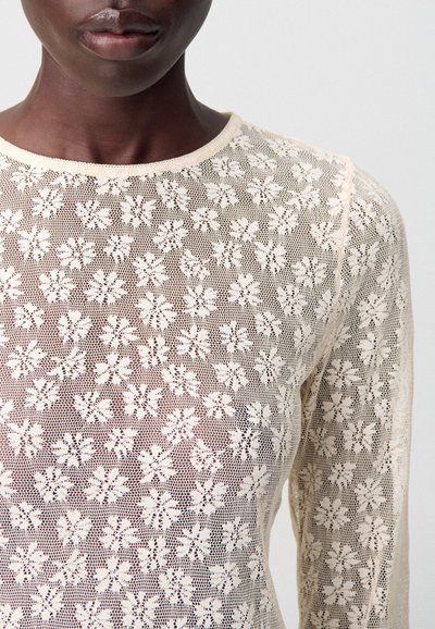 Persona de piel oscura que lleva una blusa de encaje beige transparente con patrones florales, mostrando la parte inferior del rostro desde la nariz hacia abajo y la parte superior del torso.