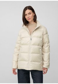 Žena v béžovej pufovacej bunde cez svetlohnedý sveter a tmavé džínsy, stojaca pred jednofarebným svetlošedým pozadím.