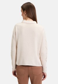 Ljus beige tröja med en texturerad yta, som har en ribbad krage och mönstrad detaljer längs ärmarna. Avskuren nederkant.