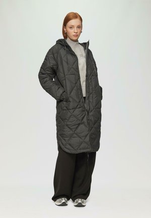 Gequilted zwarte lange jas met een capuchon, voorzien van zijzakken en een rits aan de voorkant, gecombineerd met losse zwarte broek en zilveren sneakers.
