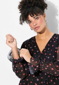 Schwarzes durchsichtiges Kleid mit einem Herzmuster in Rot und Weiß. V-Ausschnitt und gepuffte Ärmel mit elastischen Bündchen, die Textur und Detail hinzufügen.