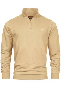 Pull à col zippé beige avec des manches longues, présentant des poignets et un ourlet côtelés. Texture douce et discret logo sur la poitrine.