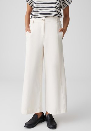 Person wearing white wide-leg pants, black loafers, and a short-sleeve striped top, hands in pockets against plain background.