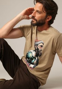 T-shirt en coton beige avec un graphique d'un homme barbu portant des lunettes de soleil et un bonnet, avec des accents verts et noirs, et du texte rouge en dessous.
