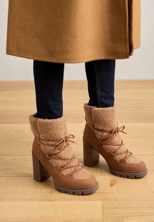 Bottes chevilles en peau retournée marron avec talons épais portées avec un jean foncé et un manteau couleur camel sur un sol en bois.