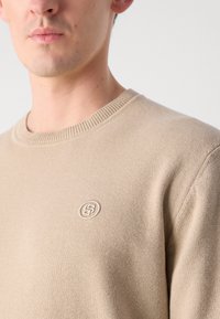Beige kötött pulóver kerek nyakkal. Texturált anyaga és diszkrét hímzett logója van a mellkasán. Puha, könnyű anyag.