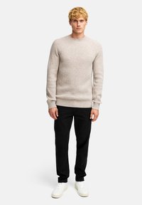 Beiger gerippter Pullover mit Rundhalsausschnitt, getragen über schwarzen Hosen und weißen Sneakern. Der Pullover hat ein strukturiertes Strickmuster und eng anliegende Ärmel.