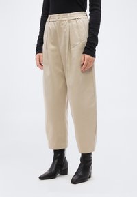 Γυναίκα με μπεζ ψηλόμεσο παντελόνι με πιέτες και μαύρα μποτάκια με τακούνι, στέκεται με χαλαρά τα χέρια στο πλάι.