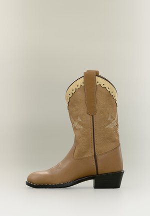 Cizme de cowboy din piele de culoare tabac, cu margine crestată crem, broderie cu pasăre și toc mic negru pe un fundal neutru.