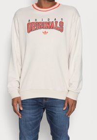 Sudadera beige claro con cuello acanalado naranja. Presenta un gran texto "Adidas Originals" en rojo. Ajuste holgado con mangas largas y dobladillo recto.
