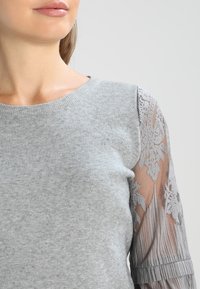 Haut en maille gris clair avec manches en dentelle aux trois-quarts, présentant des motifs floraux en dentelle élaborés et un col rond côtelé.