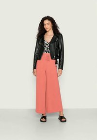 Jaqueta de couro preta, blusa com padrão floral e calças corais largas. Sandálias pretas completam o visual. Cabelo encaracolado adiciona textura.