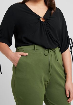Vrouw draagt een zwarte top met een sleutelgatdetail aan de voorkant en groene, hooggetailleerde broek met zijzakken en een taillekoord.