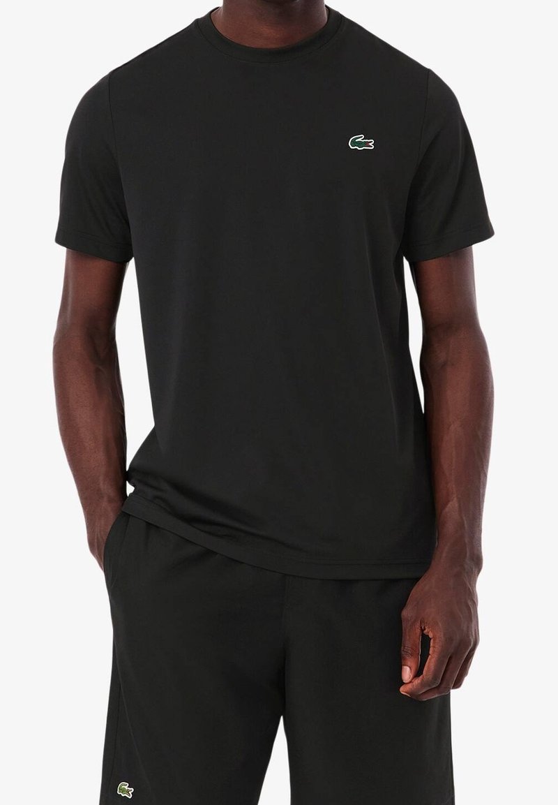 Man som bär en svart kortärmad t-shirt och shorts med en liten grön krokodil-logotyp på båda plaggen, med avslappnade händer vid sidorna.