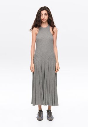 Mujer con cabello largo y rizado, que lleva un vestido gris sin mangas y zapatillas grises, situada frente a un fondo blanco.