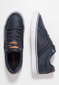 Baskets en cuir bleu marine avec semelle en caoutchouc blanche, bandes latérales texturées et accents marron sur le talon et la languette. Design à lacets.