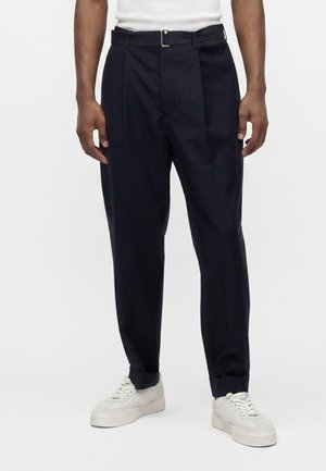 Homme portant un pantalon noir taille haute avec une ceinture assortie et des baskets blanches, debout sur un fond clair uni.
