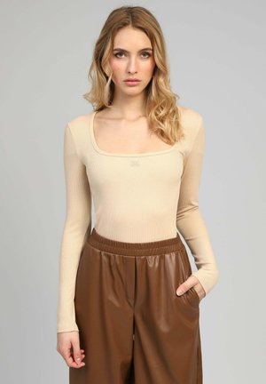 Femme blonde aux cheveux ondulés portant un haut beige à manches longues et un pantalon en cuir marron taille haute, debout avec une main dans la poche.