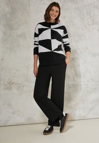 Maglione geometrico nero e grigio con orlo a coste, abbinato a pantaloni neri larghi e sneakers nere con dettagli chiari.