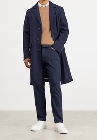 Marinblå kappa med stora knappar, beige ribbad tröja och marinblå byxor. Vita sneakers fulländar outfiten. Slätt tyg, skräddarsydd passform.