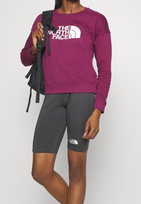 Violetti huppari, jossa on valkoinen logo etupuolella, yhdistettynä tummanharmaisiin shortseihin, joissa on sama logo. Musta reppu pidetään vasemmassa kädessä.