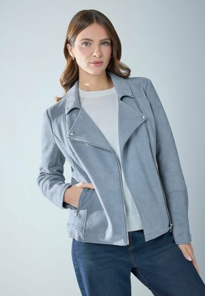 Donna con capelli castano chiaro che indossa una giacca in camoscio azzurro chiaro sopra un maglione bianco e jeans blu scuro, in posa davanti a uno sfondo neutro.