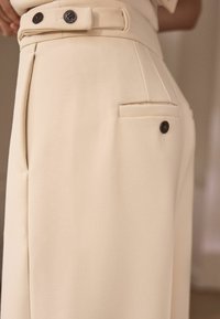 Pantaloni bej, croiți, cu o textură netedă, având o talie înaltă, două nasturi și o pocheta pe spate cu un accent de nasture închis.