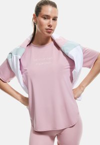 Top atlético de manga corta en color rosa con el texto "sé tu propia inspiración", complementado por una chaqueta multicolor pastel clara que descansa sobre los hombros.