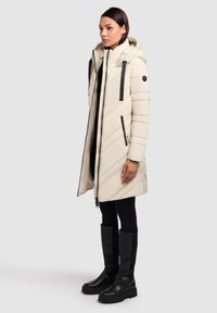 Cappotto invernale imbottito crema con cappuccio, trapuntatura diagonale, cerniere nere e dettagli neri. Indossato sopra leggings neri e stivali neri alti fino al ginocchio.
