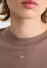 Barna pamut póló kerek nyakkal, amelynek mellkasán egy diszkrét, hímzett Nike logó található, amely megegyező árnyalatú. Sima textúra.