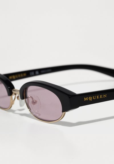 Mustaga raami prillid ümmarguste lilla objektiivide ja kuldsete detailidega, kus templitel on kuldsete tähtedega kirjutatud "McQueen".