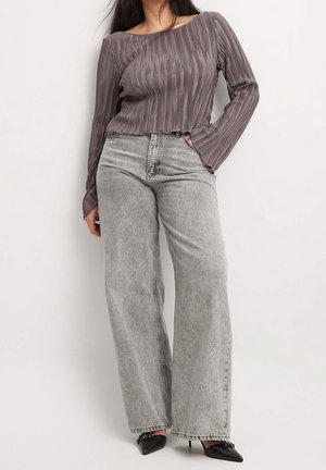 Woman wearing a pleated long-sleeve grayish-brown top, high-waisted light gray wide-leg jeans, and black pointed heels.