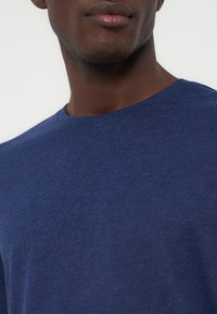 Κοντινό πλάνο ενός ατόμου που φοράει μια απλή μπλε μπλούζα με στρογγυλή λαιμόκοψη, δείχνοντας τον λαιμό και το κάτω μέρος του προσώπου σε απλό φόντο.