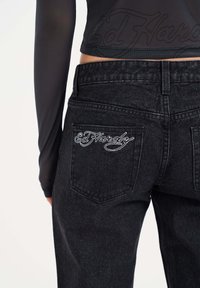 Jeans de mezclilla negra con el logo blanco bordado de "Ed Hardy" en el bolsillo trasero. La tela parece texturizada y ligeramente desgastada.