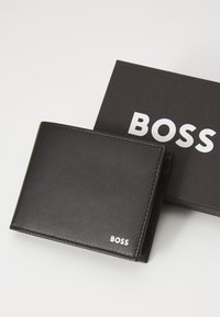 Portafoglio in pelle nera con logo in argento. Presenta una texture liscia e cuciture precise, confezionato in una scatola nera con testo bianco in grassetto.