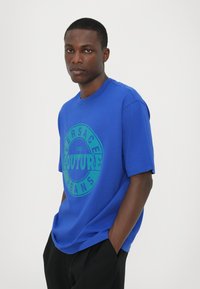 Mėlynas trumpas rankovėmis t-shirt, pagamintas iš medvilnės, su apvaliu grafiniu motyvu, kuriame yra žaliaisiais „VERSACE COUTURE JEANS“ tekstas. Įprastas siluetas.