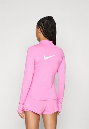 Femme avec des cheveux tressés portant un haut de sport à manches longues rose et un short assorti, de dos, montrant un logo Nike blanc sur le dos.