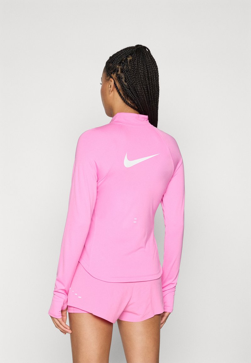 Frau mit geflochtenem Haar, die ein pinkfarbenes langärmliges Sportoberteil und passende Shorts trägt, mit dem Rücken zur Kamera gewandt. Auf dem Rücken ist ein weißes Nike-Logo zu sehen.