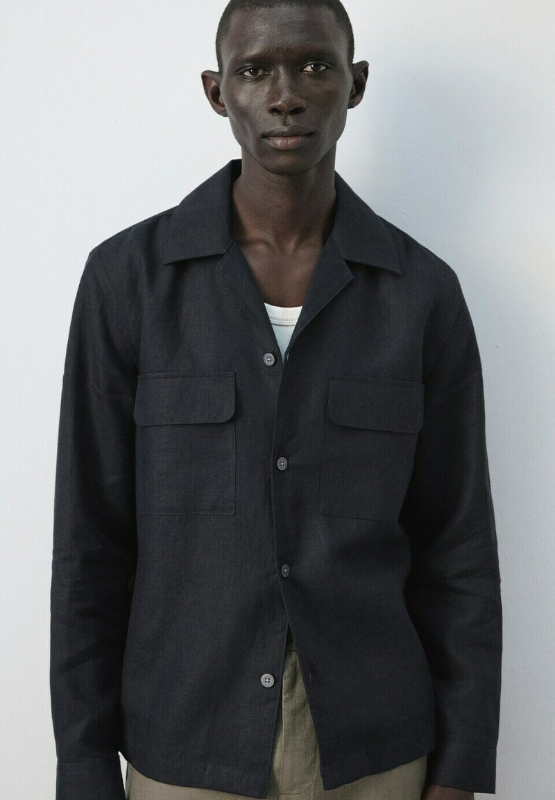 Jeune homme à la peau foncée portant une veste boutonnée foncée par-dessus une chemise blanche, sur un fond clair uni.