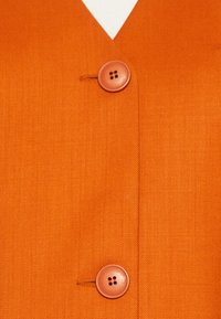 Oranžový oděv s texturovanou tkaninou, V-výstřihem a dvěma kulatými knoflíky. Knoflíky jsou světle oranžové se čtyřmi dírkami.