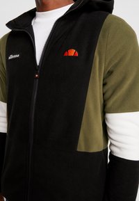 Giacca in felpa con zip caratterizzata da pannelli neri, verde oliva e bianchi. Include logo ricamato, colletto alto e tasche laterali.