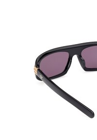 Solen glasögon med en svart plastbåge, lila tonade linser och guld accenter på skalmarna, med en logotypdetalj nära gångjärnet.