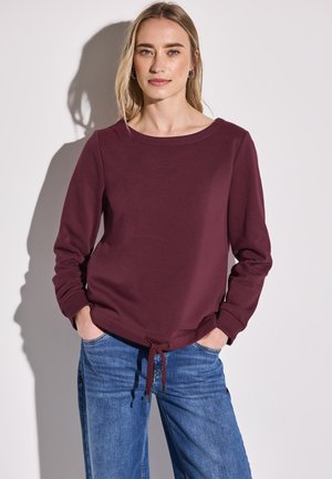 Bordeaux sweatshirt met lange mouwen, ronde halslijn en verstelbare trekkoord aan de onderrand. Gecombineerd met lichtblauwe wijde jeans.