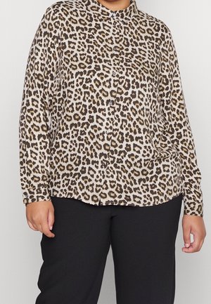 Osoba nosząca długą koszulę w panterkę zapinaną na guziki i czarne spodnie, stojąca na neutralnym tle.