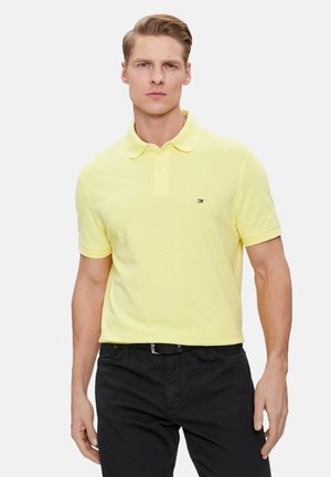 Camisa polo amarilla de algodón, con cuello clásico, mangas cortas y un pequeño logotipo en el lado izquierdo del pecho, combinada con pantalones negros.