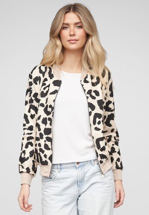 Kobieta o długich blond włosach, ubrana w beżowo-czarną kurtkę na suwak w panterkę, na białej koszulce i jasnoniebieskich dżinsach.