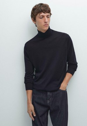 Maglione a collo alto nero, morbido al tatto, design aderente, maniche lunghe, polsini a coste, abbinato a jeans scuri, su uno sfondo chiaro.