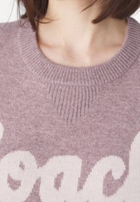 Maglione color malva chiaro con scollo rotondo a coste e dettaglio testurizzato al colletto; presenta un logo bianco in un carattere giocoso sulla parte anteriore.