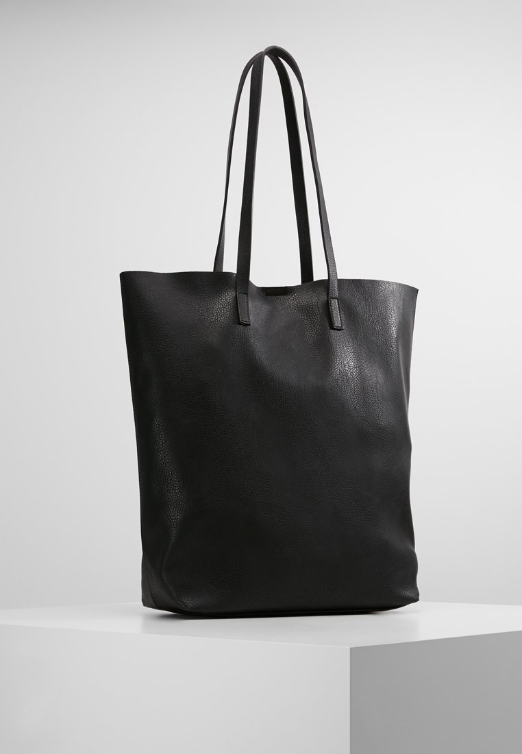 Sac cabas en cuir noir avec longues anses, debout sur un piédestal blanc sur fond uni gris clair.